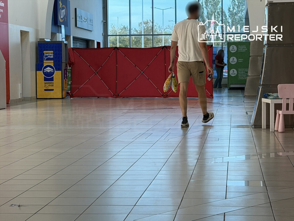 Mężczyzna w krótkich spodniach przechodzi przez przestronny hol z czerwonym parawanem w tle.
