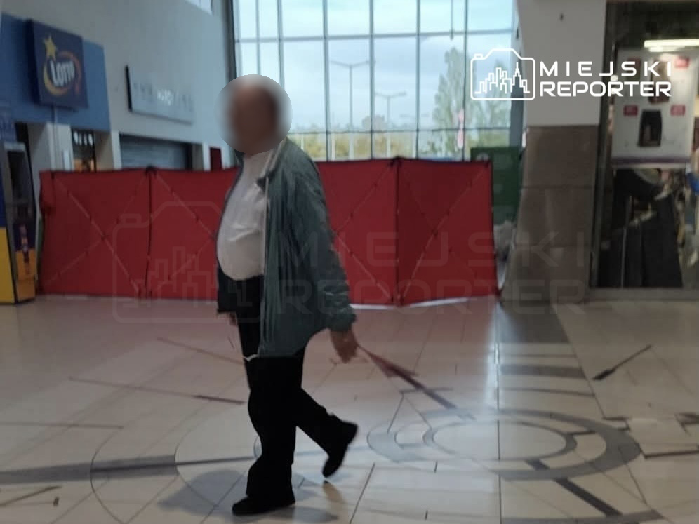 Mężczyzna w niebieskiej kurtce spacerujący po centrum handlowym, w tle czerwone przesłony.