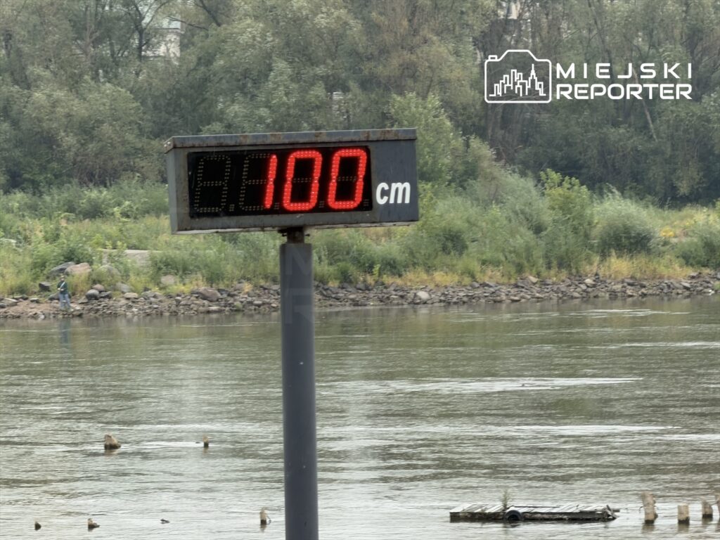Wyświetlacz z cyframi 100 cm wskazującymi poziom wody w rzece Wisła, w tle wędkarz na brzegu.