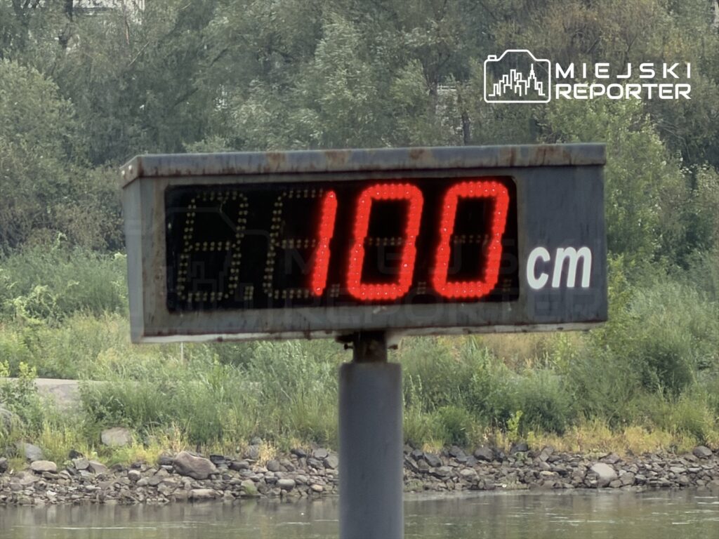 Wskaźnik poziomu wody pokazujący 100 cm, w tle zieleń i brzeg rzeki Wisły.