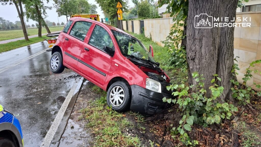 Czerwony samochód osobowy wjechał w pień drzewa na poboczu drogi, obok widoczny pojazd służb ratunkowych.