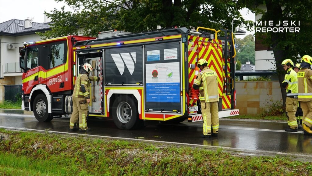 Strażacy w mundurach rozmawiają przy wozie strażackim na mokrej drodze, obok nowoczesnych budynków mieszkalnych.