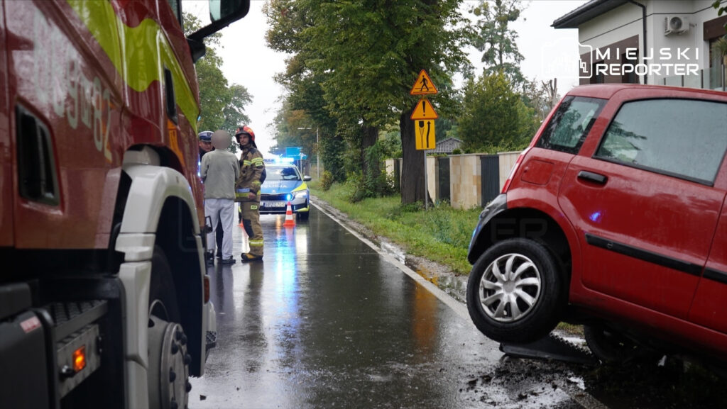 Na mokrej drodze stoi czerwona osobówka z uniesionym kołem, obok grupka ratowników i policjantów w mundurach.