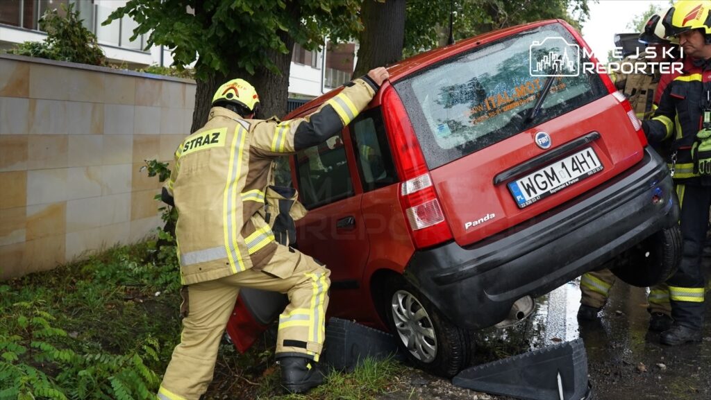 Strażacy podnoszą czerwoną fiata Pandę, która wjechała w drzewo, w otoczeniu zieleni i budynków.