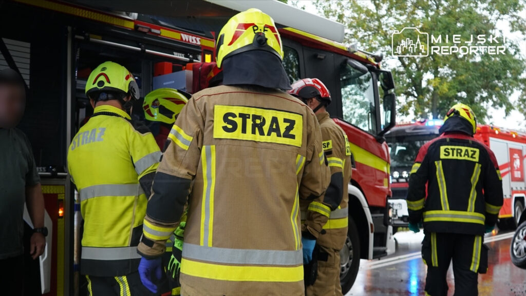 Grupa strażaków w specjalistycznych strojach omawia sytuację obok wozu strażackiego na miejscu wypadku.