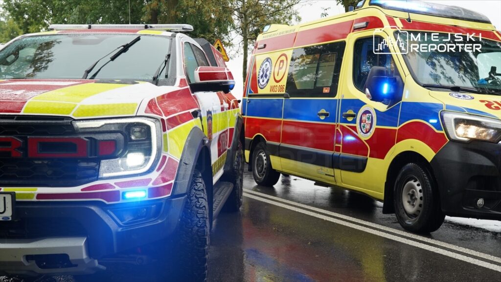 Pojazdy ratunkowe, w tym radiowóz i karetka, zatrzymane na deszczowej drodze, z włączonymi sygnałami świetlnymi.
