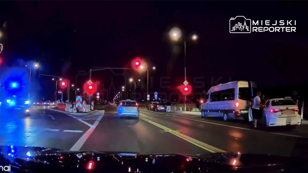 Niebieski samochód zatrzymuje się na czerwonym świetle, obok widoczny biały pojazd z pasażerami, w tle widać policyjny radiowóz.