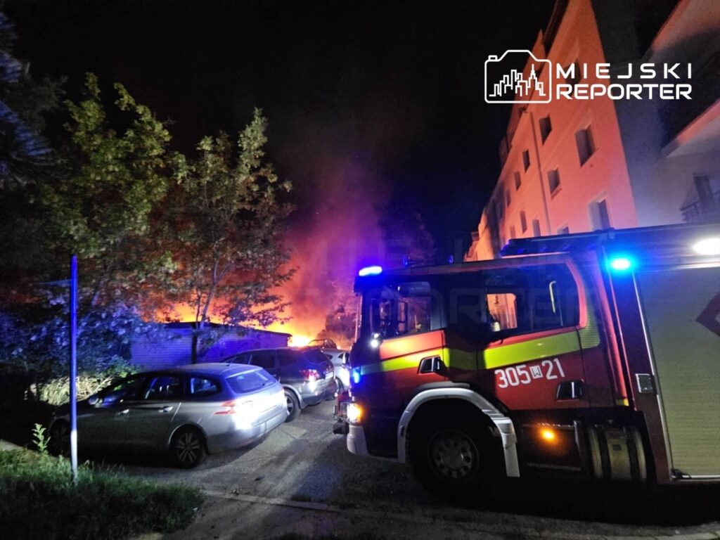 W nocy widać ogień wydobywający się z altany, obok zaparkowane samochody oraz woz strażacki z włączonymi sygnałami świetlnymi.