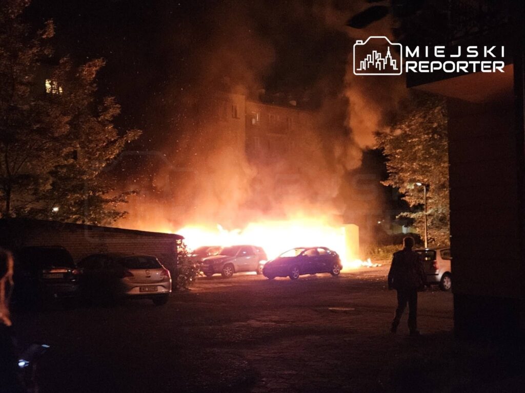 Płonąca altana i kilka samochodów w nocy na osiedlowym parkingu, w tle widać dym i ogień.