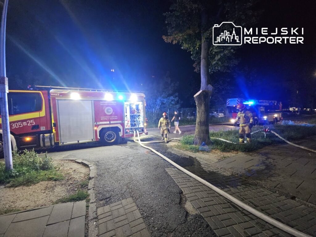Strażacy w mundurach przeciwpożarowych pracują przy wozie strażackim, rozkładając węże na oświetlonym nocą terenie.