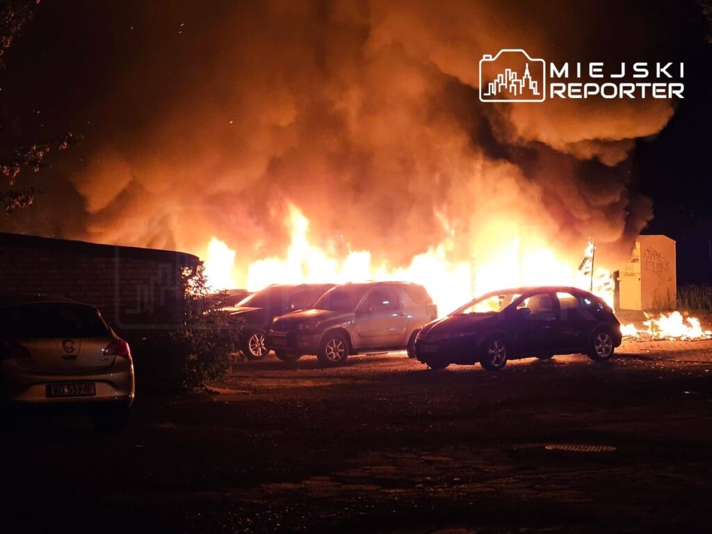 Płonąca altana i zaparkowane samochody otoczone intensywnym ogniem i dymem w ciemnej scenerii.