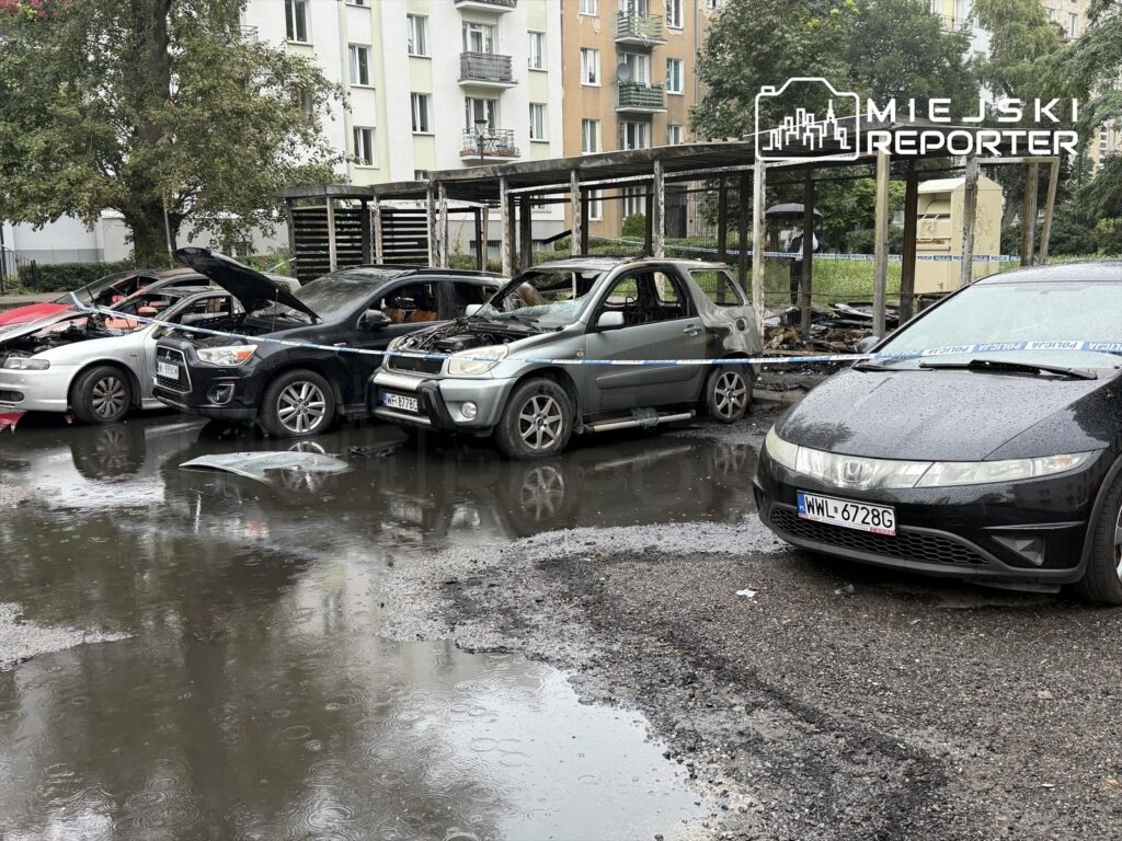 Zniszczone samochody z otwartymi maskami stoją na mokrej nawierzchni, w tle widoczna altana oraz budynki mieszkalne.