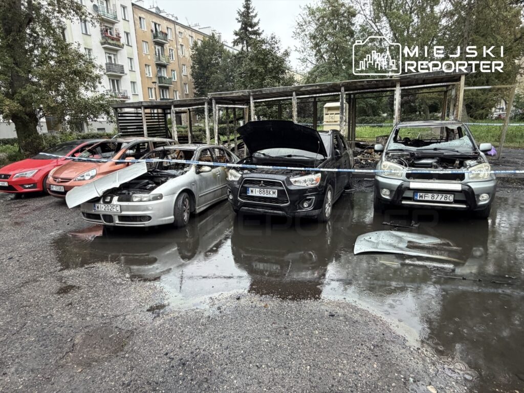 Zniszczone samochody stoją na mokrym placu, obok spalonej altany, w tle widać bloki mieszkalne.