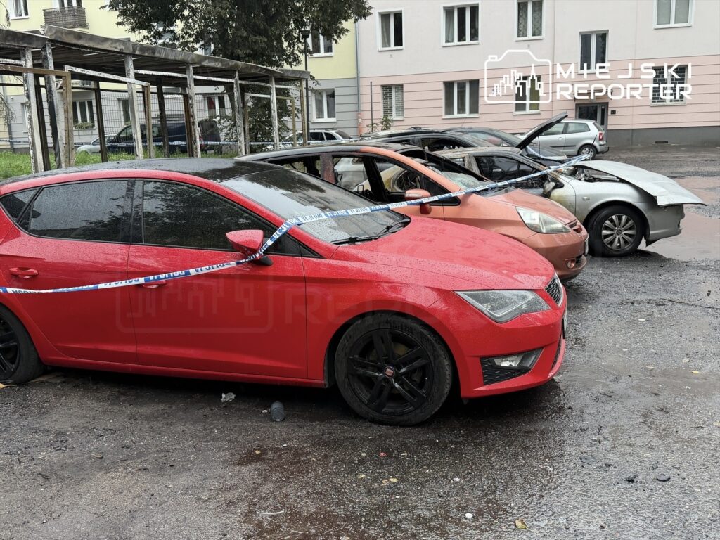 Czerwony samochód osobowy z czarnymi felgami stoi na mokrej nawierzchni obok uszkodzonych pojazdów, otoczony taśmą policyjną.