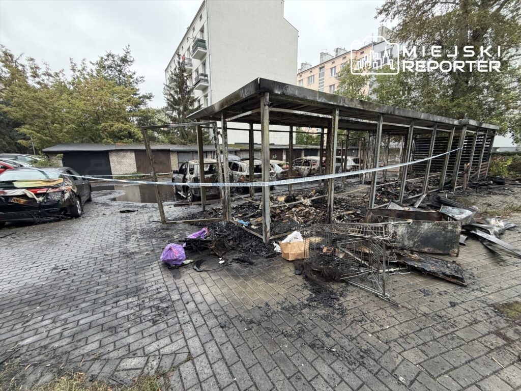 Zniszczona altana i spalone samochody na parkingu, w tle budynki mieszkalne i drzewa. Wokół rozrzucone resztki pożaru.