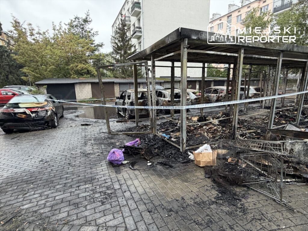 Zniszczona altana i spalone samochody w parkingu, otoczone czarnym popiołem i resztkami pożaru.