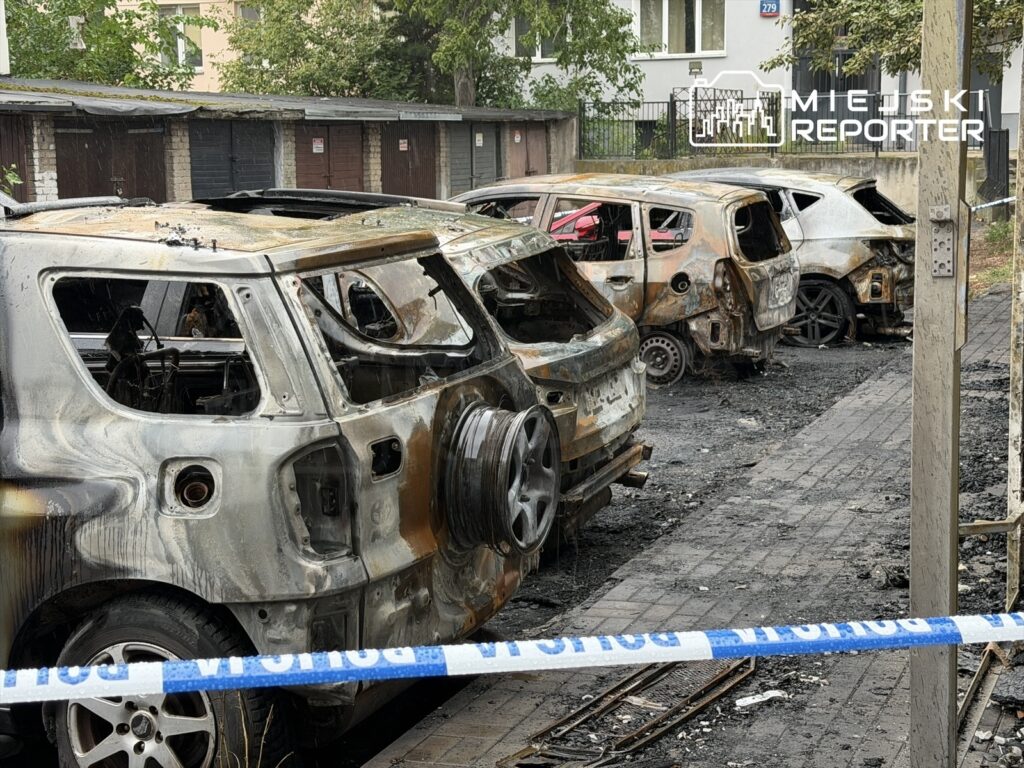 Zniszczone, spalone samochody stoją na czarnym asfalcie, otoczone taśmą policyjną w zniszczonym otoczeniu.