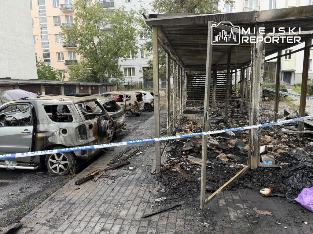 Cztery spalone samochody stoją na zniszczonym parkingu, obok spalonej altany, otoczone taśmą zabezpieczającą.