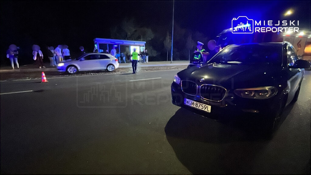 Grupa osób stoi przy przystanku autobusowym, a w tle widoczny jest radiowóz policyjny oraz samochód osobowy.