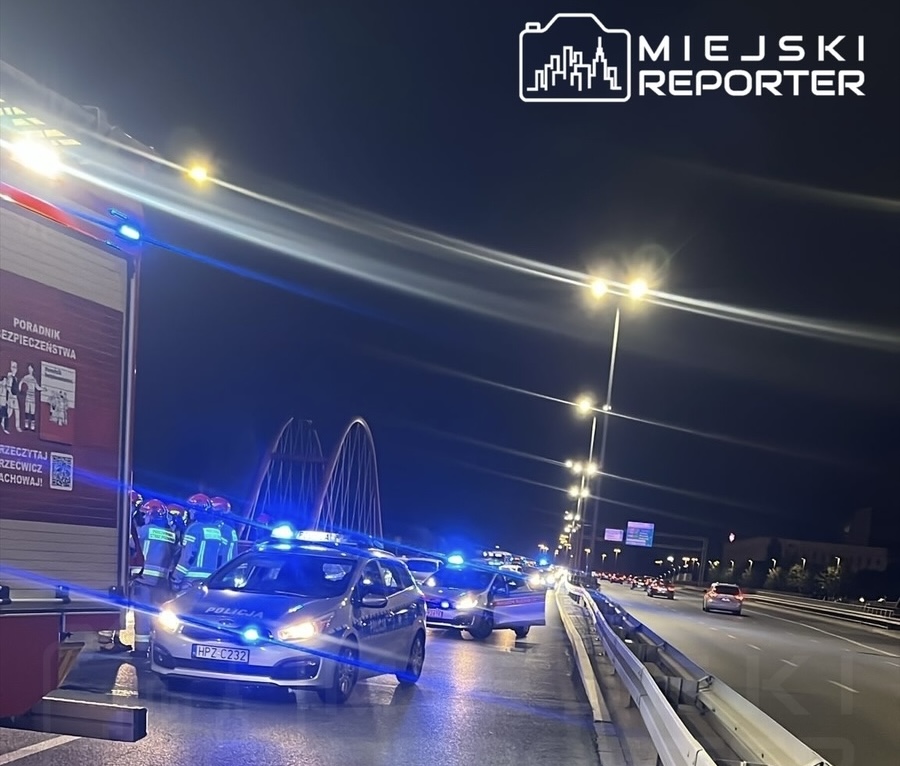 Na drodze w nocy zespół ratunkowy i policja zabezpieczają miejsce wypadku, obok zaparkowane radiowozy z włączonymi sygnałami świetlnymi.