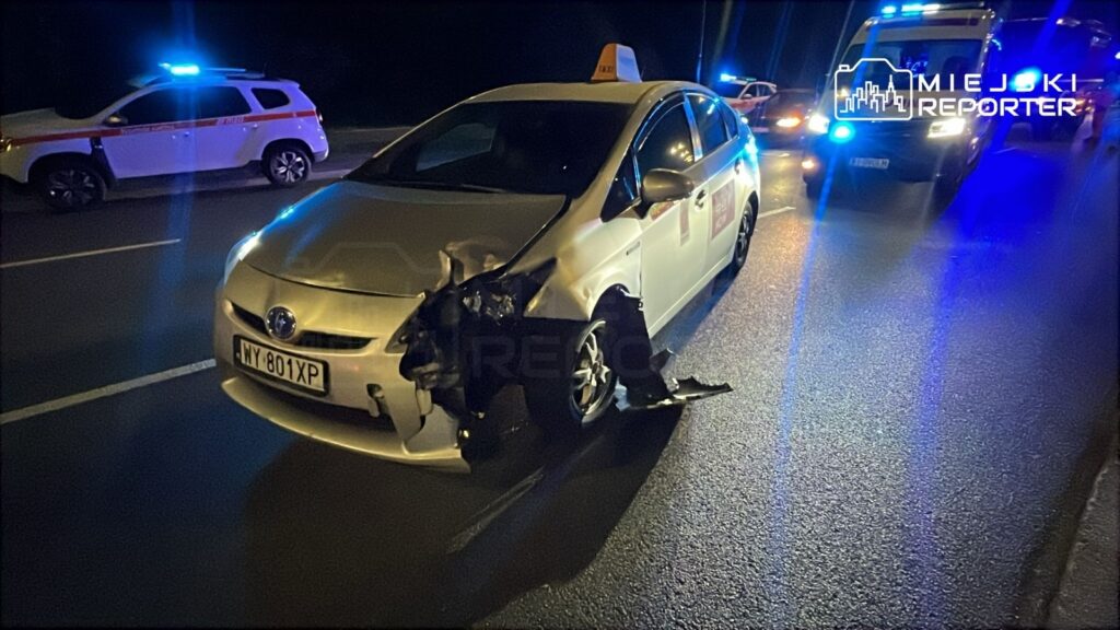 Uszkodzony samochód osobowy z oznaczeniem taxi stoi na drodze, obok niego widać pojazdy służb ratunkowych z włączonymi sygnałami świetlnymi.