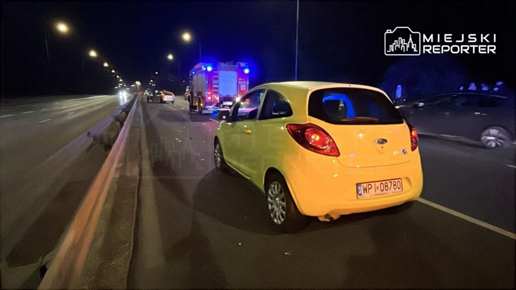 Żółty samochód osobowy stoi na poboczu drogi, obok niego widać pojazd straży pożarnej z włączonymi sygnałami świetlnymi.