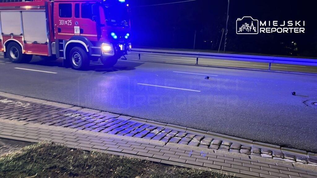 Czerwony wóz strażacki z niebieskimi sygnałami stoi na oświetlonej ulicy obok pasów ruchu, w tle widać krawężnik.