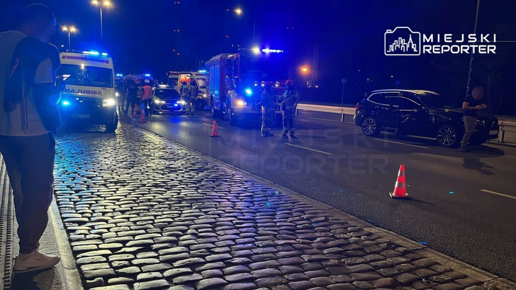Na oświetlonej ulicy stoi ambulans oraz radiowóz, obok grupka strażaków w mundurach. W tle widać osobowe auto z włączonymi światłami awaryjnymi.
