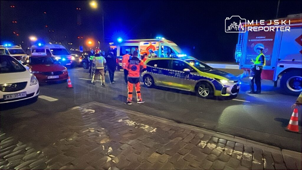 Na drodze, w nocy, zespół ratunkowy oraz policja zabezpieczają miejsce wypadku, obok stoją zaparkowane pojazdy.