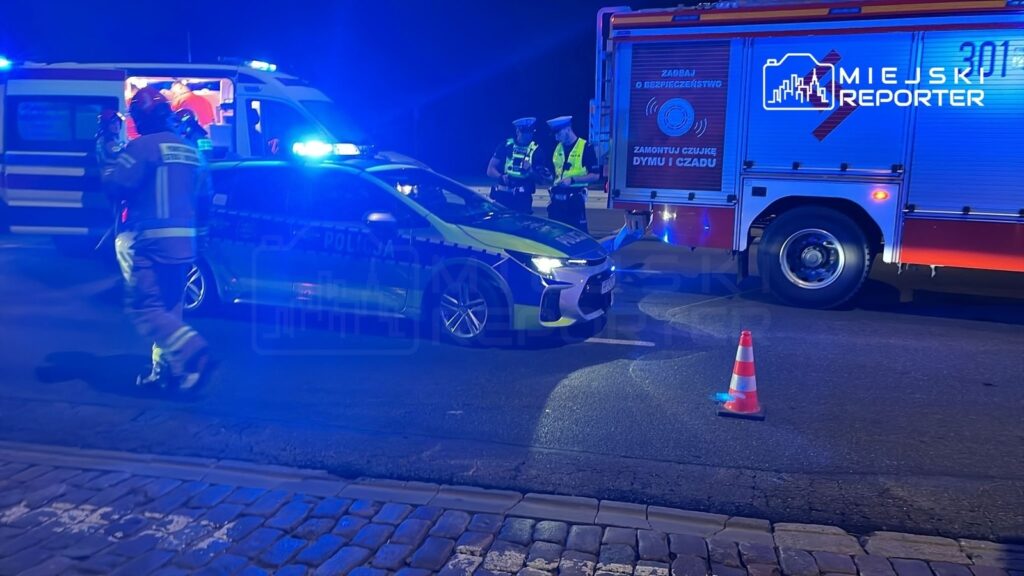 Na drodze stoi radiowóz policyjny z włączonymi sygnałami świetlnymi, obok pracują strażacy i policjanci.
