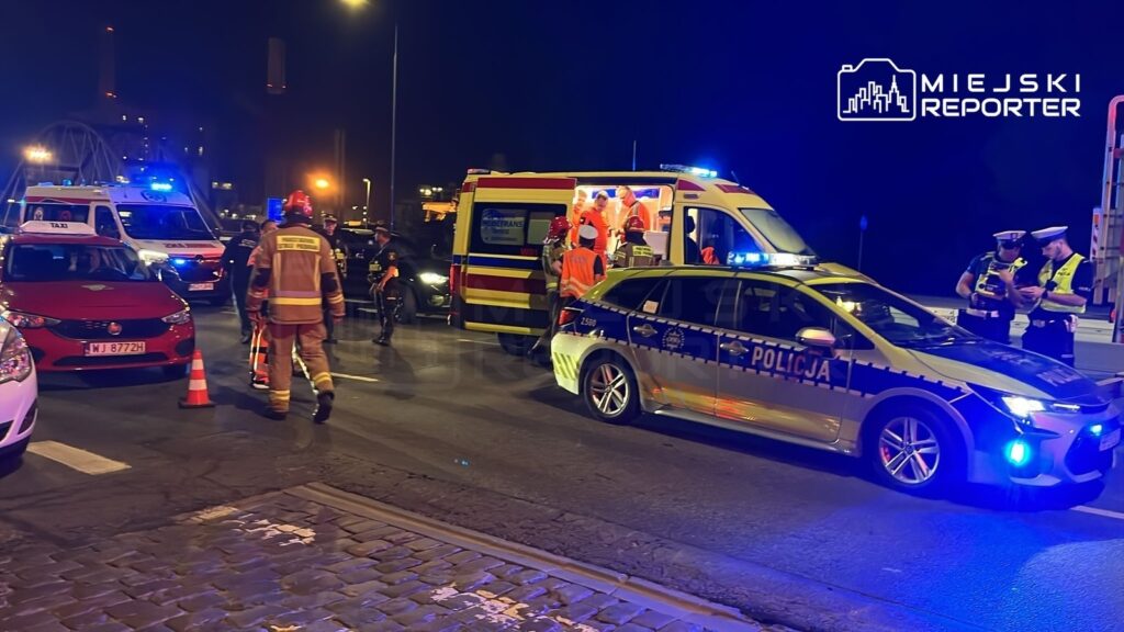 Na drodze nocą, w miejscu wypadku, policjanci i strażacy udzielają pomocy, a karetka czeka na rannych.