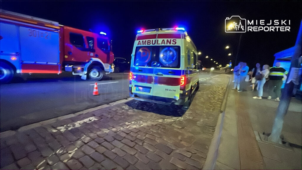 Ambulans z włączonymi sygnałami świetlnymi stoi na drodze, obok grupy ludzi i wozu strażackiego.