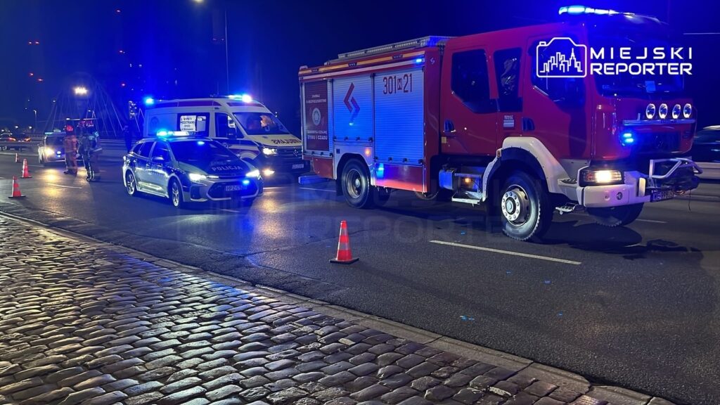 Na drodze zaparkowane są pojazdy służb ratunkowych, w tym wóz strażacki i radiowóz, a obok stoją funkcjonariusze w mundurach.
