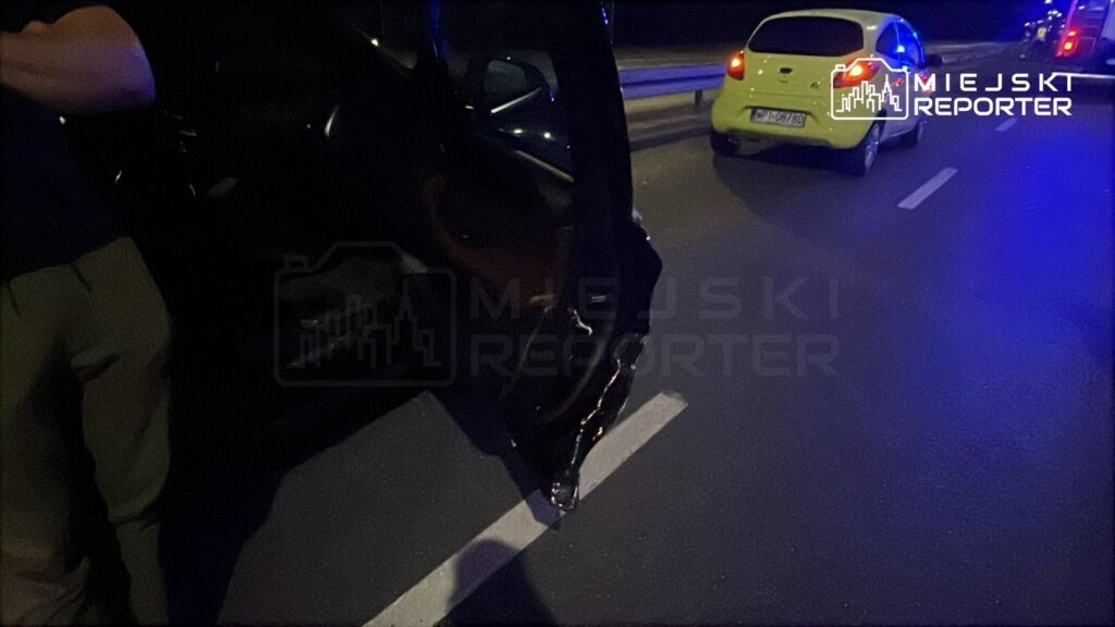 Mężczyzna w ciemnym ubraniu stoi obok otwartego drzwi samochodu na drodze, w tle widać zaparkowany żółty pojazd.