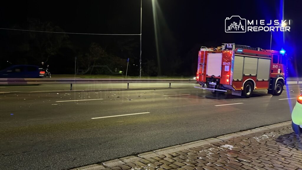 Czerwony wóz strażacki z włączonymi sygnałami świetlnymi przejeżdża obok uszkodzonego mienia na drodze nocą.