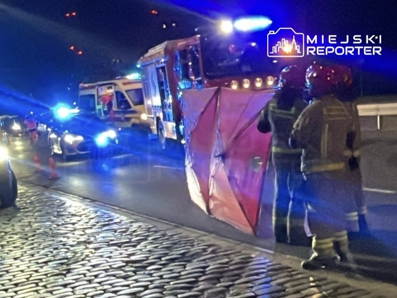 Strażacy w hełmach i mundurach zabezpieczają miejsce wypadku, obok widać samochód strażacki i pojazdy policyjne.