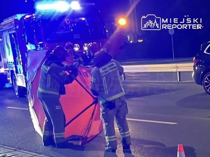 Strażacy ustawiają osłonę na drodze podczas akcji ratunkowej w nocy, obok wozu strażackiego z włączonymi sygnałami.