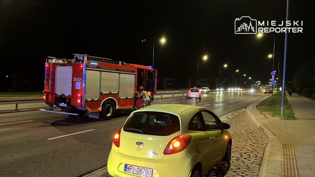 Czerwony wóz strażacki na drodze, strażak wyciągający sprzęt, w tle ruch uliczny w nocy.