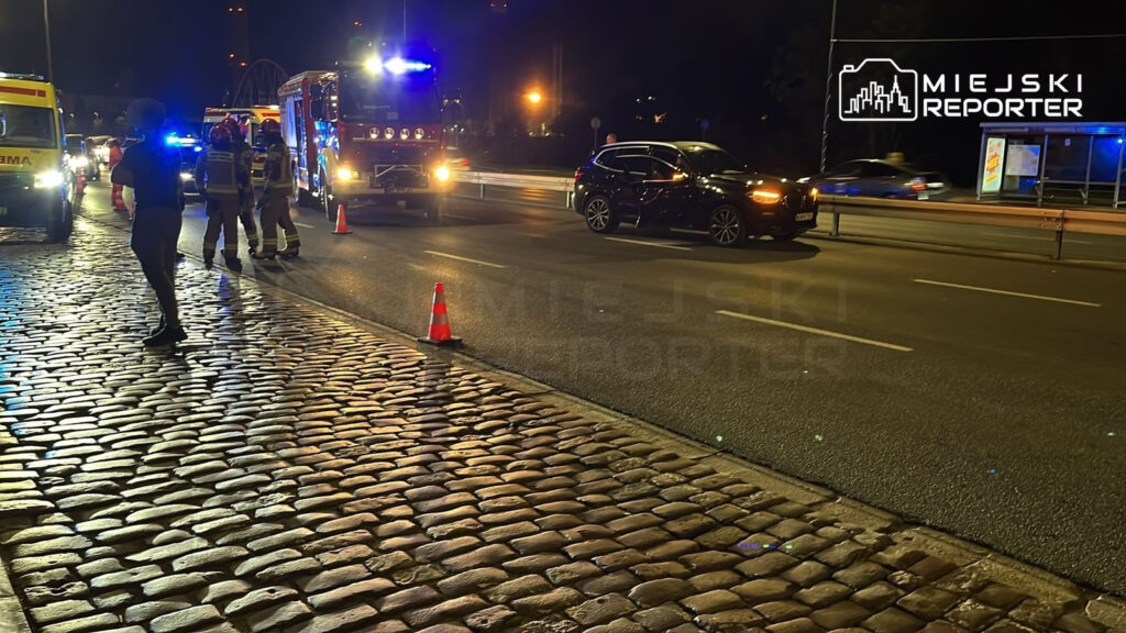 Grupa strażaków i ratowników medycznych pracuje na oświetlonej ulicy, obok stojącego pojazdu z włączonymi sygnalizatorami.