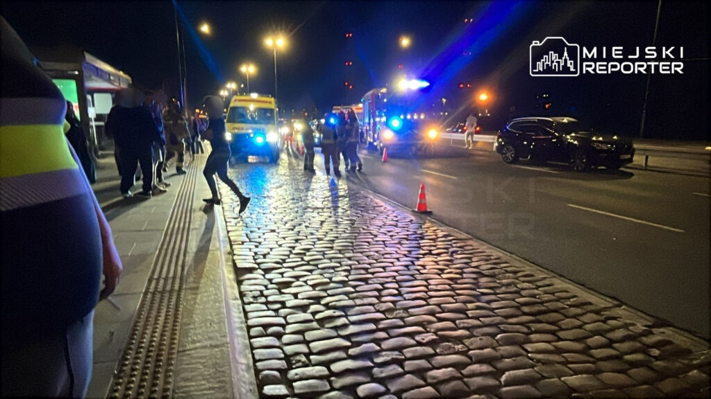 Grupa ludzi w pobliżu policyjnych i ratunkowych pojazdów na oświetlonej ulicy, w tle leżące pachołki.