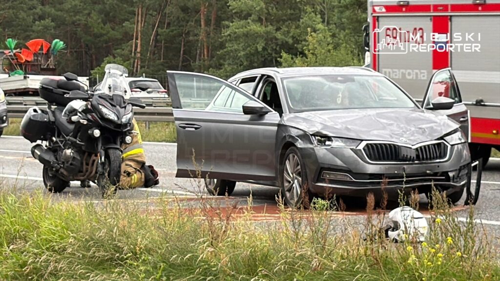 Motocyklista w mundurze klęczy obok uszkodzonego motocykla, a obok stoi rozbita szara Skoda z otwartymi drzwiami na drodze.
