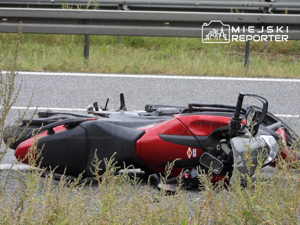 Czerwony motocykl leży na jezdni przy drodze, otoczony wysoką trawą i barierką ochronną.