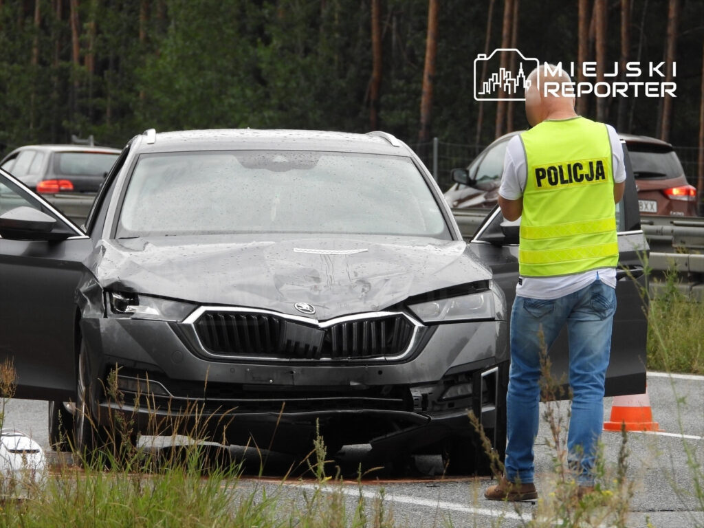 Czarny samochód osobowy z uszkodzoną przednią częścią stoi na drodze, obok policjant w kamizelce odblaskowej prowadzi czynności.