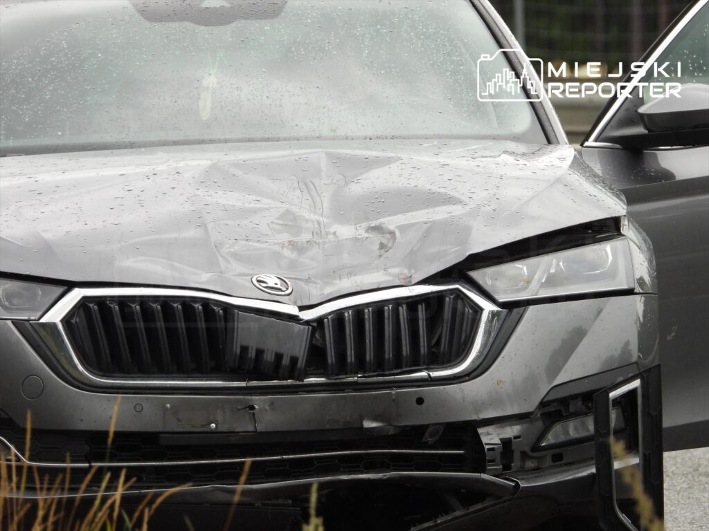 Czarny samochód osobowy z uszkodzoną maską i zderzakiem, stojący na drodze w deszczowej pogodzie.