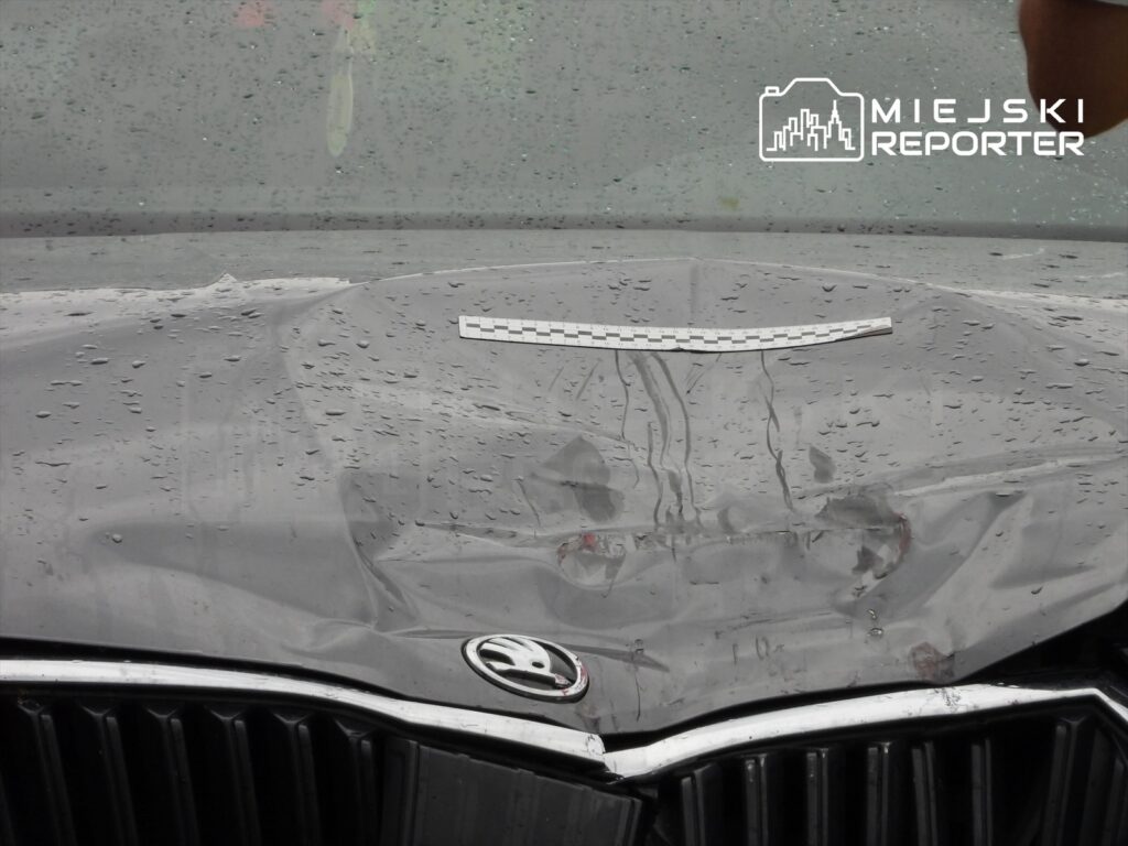 Uszkodzona maska ciemnoszarego samochodu osobowego z logo Skoda, pokryta kroplami deszczu.