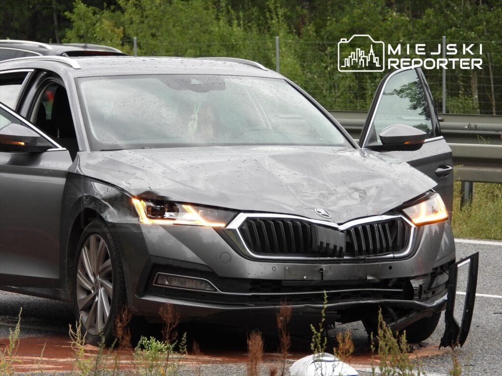 Uszkodzona szara Skoda stoi na poboczu drogi, z otwartymi drzwiami i widocznymi śladami kolizji.