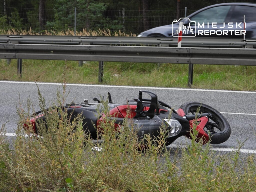 Przewrócony motocykl leży na poboczu drogi, otoczony przez wysoką trawę i krzewy. W tle widać przejeżdżający samochód.