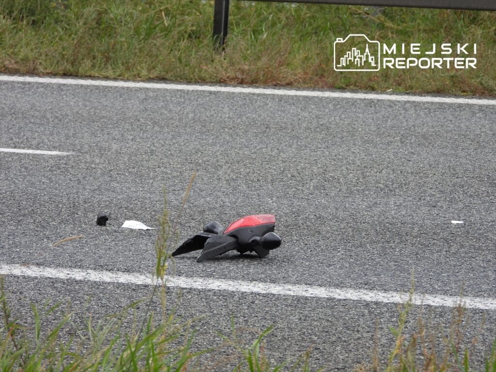 Czarna część motocykla leży na drodze, obok znajduje się fragment czerwonego elementu, otoczenie to trawa.