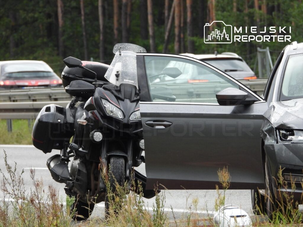 Czarny motocykl stojący obok uszkodzonego samochodu osobowego na drodze, w tle widać inne pojazdy.