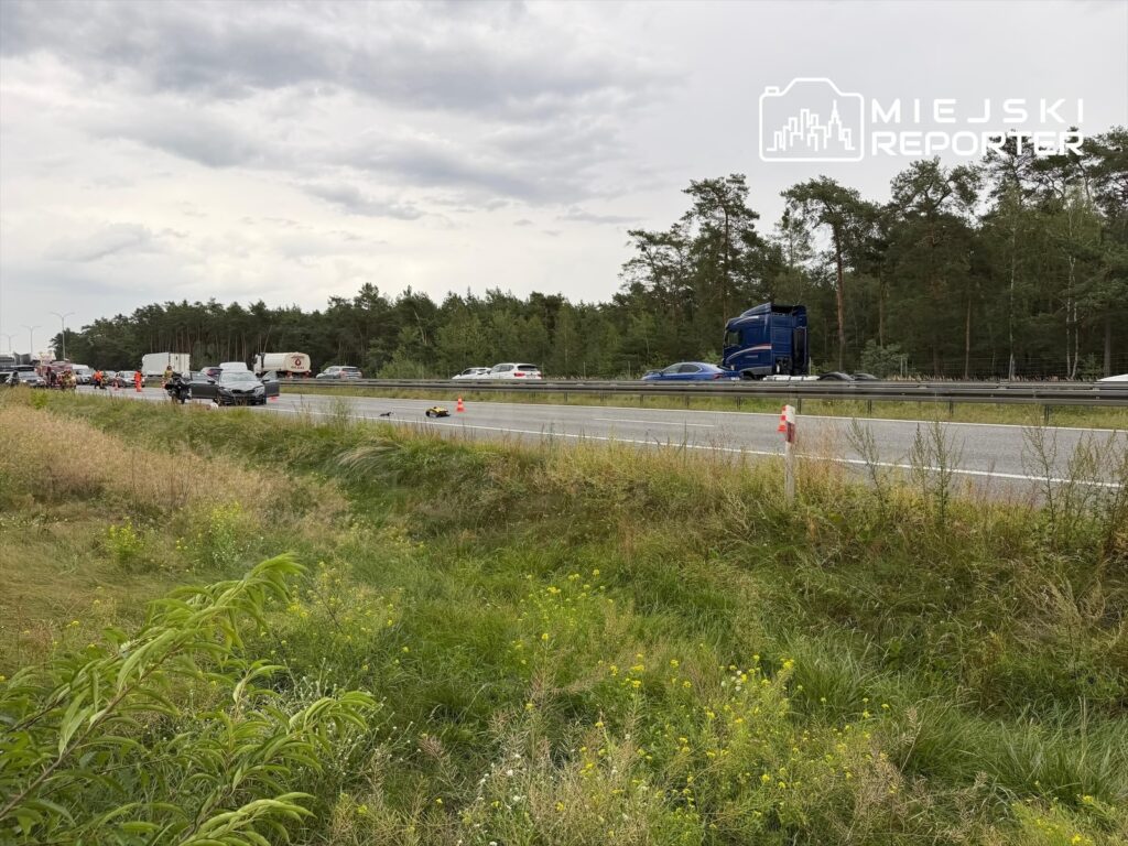 Na poboczu drogi leżą dwa motocykle, obok nich pracują służby ratunkowe, a w tle widać ruch samochodowy.